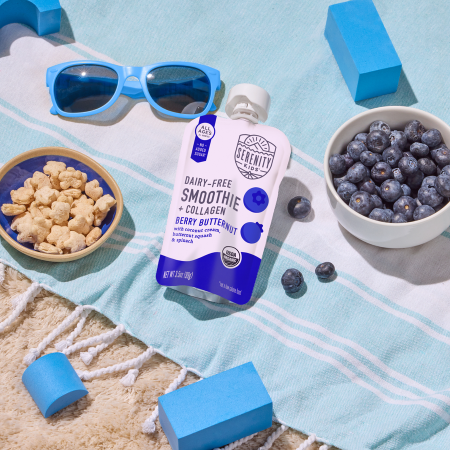 Serenity Kids smoothie pouch with blueberries, snacks, and sunglasses on a beach towel.