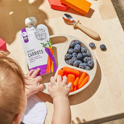 Baby reaching for a package of organic carrots next to a bowl of blueberries and carrots on a wooden table.