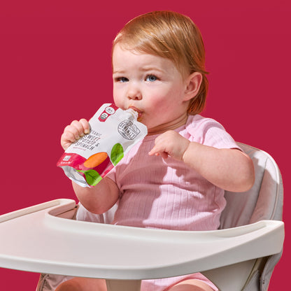 Child in a high chair holding a baby food pouch against a red background