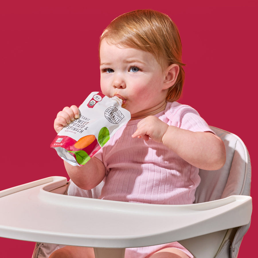 Child in a high chair holding a baby food pouch against a red background